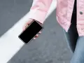 Mano sujetando un móvil apagado, con chaqueta rosa y jeans, sobre un paso de peatones.