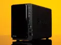 Servidor NAS Synology DS214play negro con luces indicadoras y logo en relieve, sobre fondo amarillo.