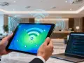 Persona usa una tablet para monitorizar red wifi, con gráficos de señal y puntos de conexión. Un portátil con código está cerca.