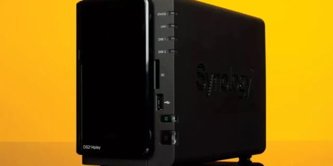 Servidor NAS Synology DS214play negro con luces indicadoras y logo en relieve, sobre fondo amarillo.