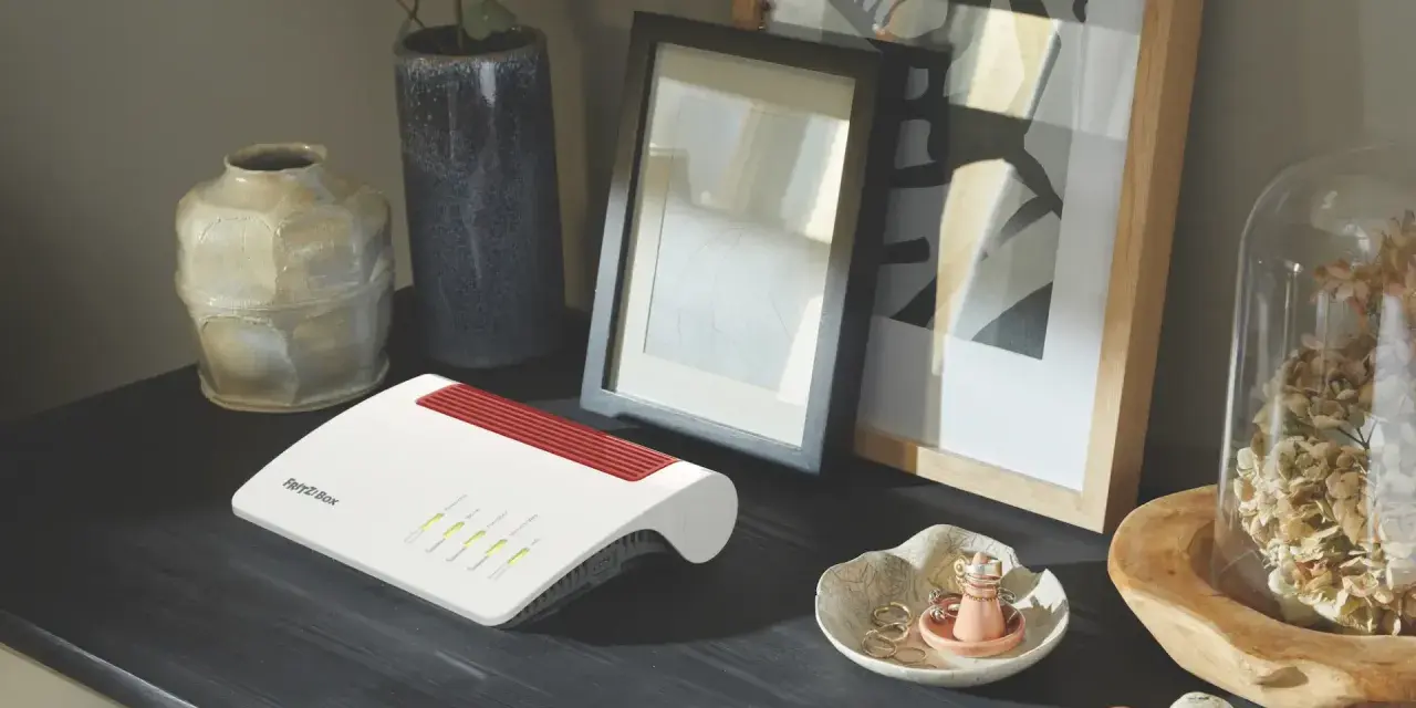 Router firewall blanco y rojo, con luces verdes encendidas, sobre un escritorio oscuro junto a jarrones y cuadros.