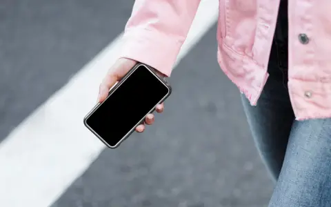 Mano sujetando un móvil apagado, con chaqueta rosa y jeans, sobre un paso de peatones.