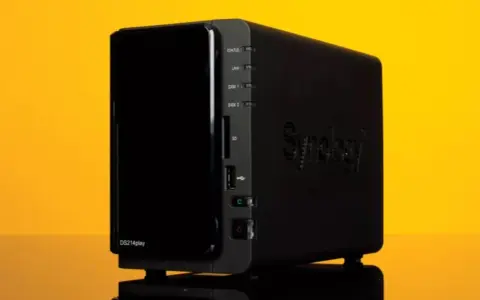Servidor NAS Synology DS214play negro con luces indicadoras y logo en relieve, sobre fondo amarillo.