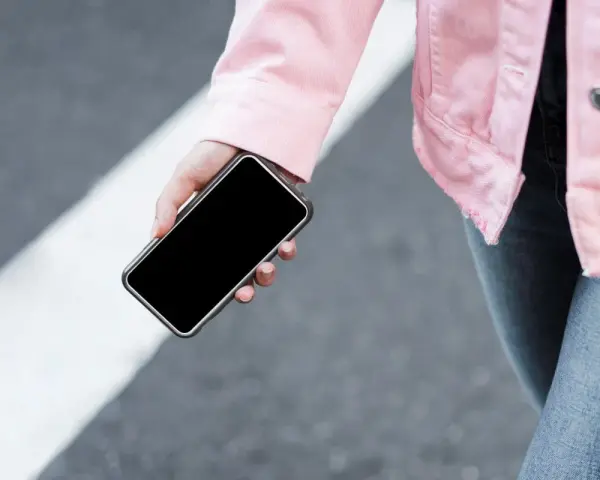 Mano sujetando un móvil apagado, con chaqueta rosa y jeans, sobre un paso de peatones.