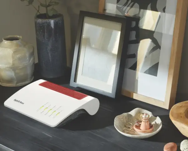 Router firewall blanco y rojo, con luces verdes encendidas, sobre un escritorio oscuro junto a jarrones y cuadros.