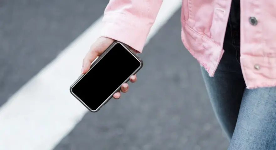 Mano sujetando un móvil apagado, con chaqueta rosa y jeans, sobre un paso de peatones.