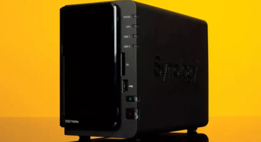Servidor NAS Synology DS214play negro con luces indicadoras y logo en relieve, sobre fondo amarillo.