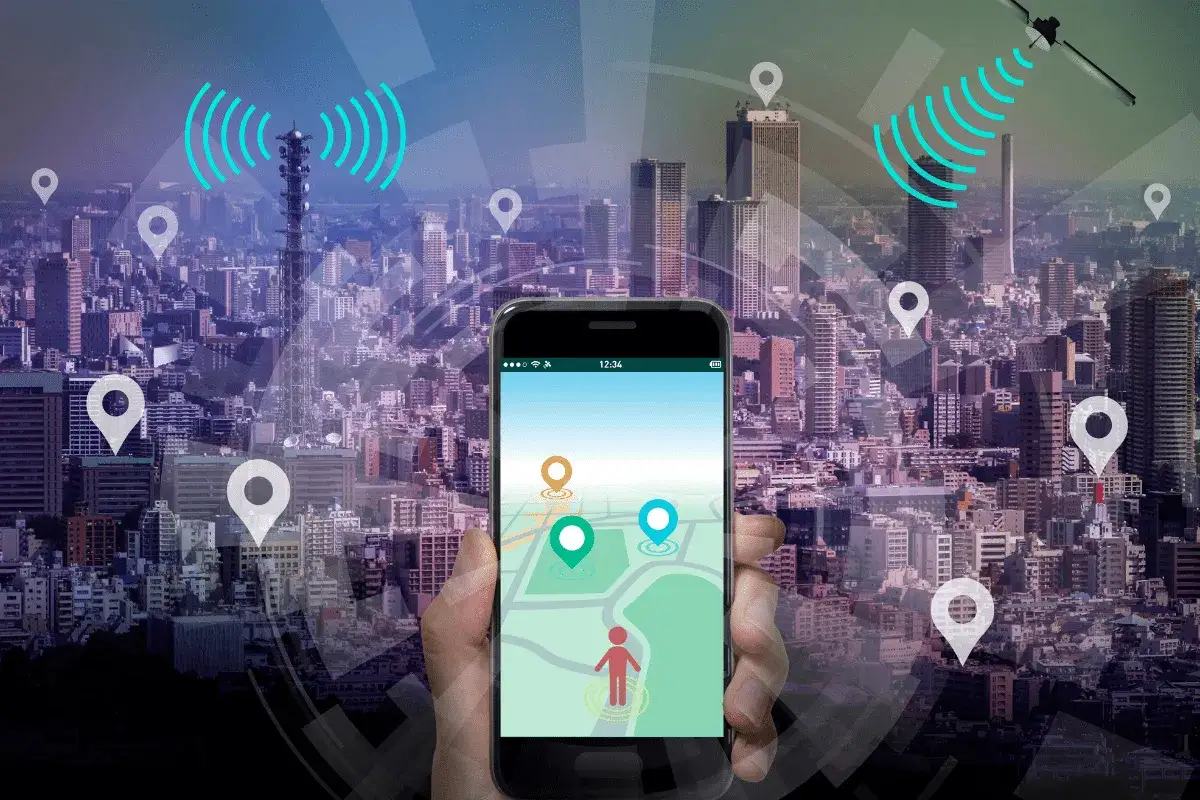 Mano sostiene móvil con mapa y pines de localización. La ciudad de fondo y antenas sugieren la tecnología para localizar persona por móvil.