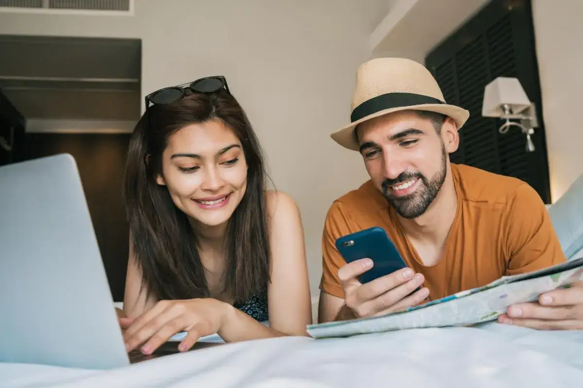 Pareja sonriente planeando su viaje, ella en el portátil y él con el móvil y un mapa. booking es fiable para organizar aventuras.